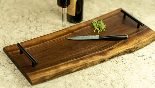 This image features a live edge serving board made from dark wood with black metal handles. A sharp knife and a sprig of rosemary are placed on the board, while a wine bottle and glass sit in the background, enhancing the rustic charm of the setting.