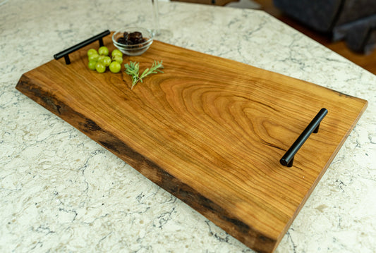 This image showcases a handcrafted live edge serving board made from rich brown wood. It features black metal handles and is adorned with green grapes, a sprig of rosemary, and a small bowl of dark preserves, resting on a marble countertop.